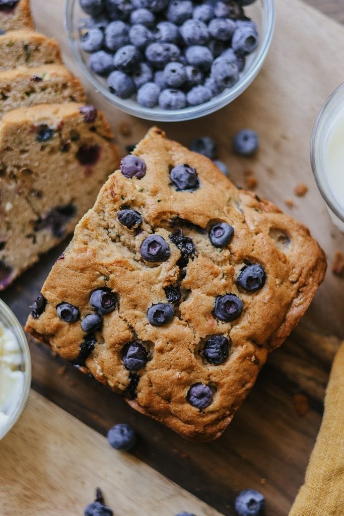 A half loaf of zucchini blueberry bread on a white plate with a slice cut and blueberries scattered around.