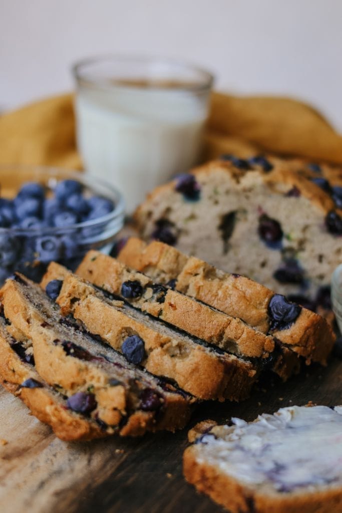 A loaf of zucchini blueberry bread with several slices cut, displayed on a wooden cutting board with blueberries scattered around.