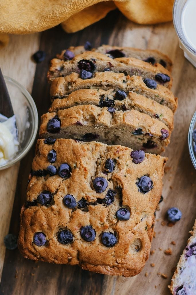 A loaf of zucchini blueberry bread partially sliced on a wooden cutting board with fresh blueberries nearby.