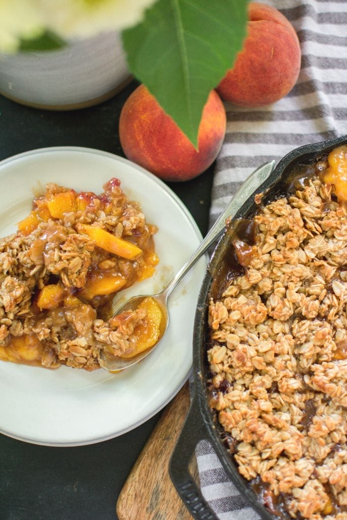 A cast iron skillet filled with a peach crisp A white plate and a spoon are beside the skillet