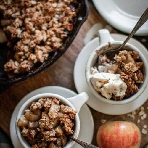 two bowls with apple crisp and vanilla ice cream. In the background is a cast iron skillet with apple crips