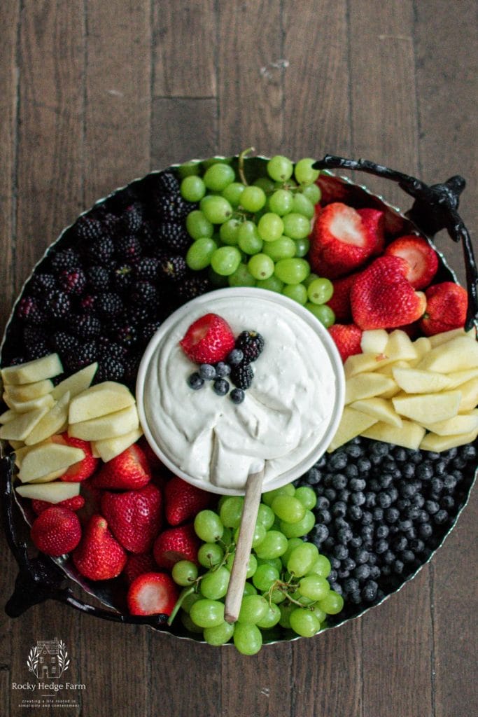 A large round tray filled with strawberries, blueberries, blackberries, apples and grapes with a white bowl in the center filled with cream cheese fruit dip. 
