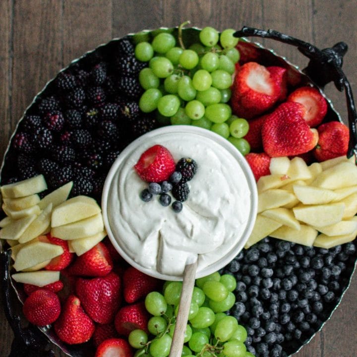 A large round tray filled with strawberries, blueberries, blackberries, apples and grapes with a white bowl in the center filled with cream cheese fruit dip.