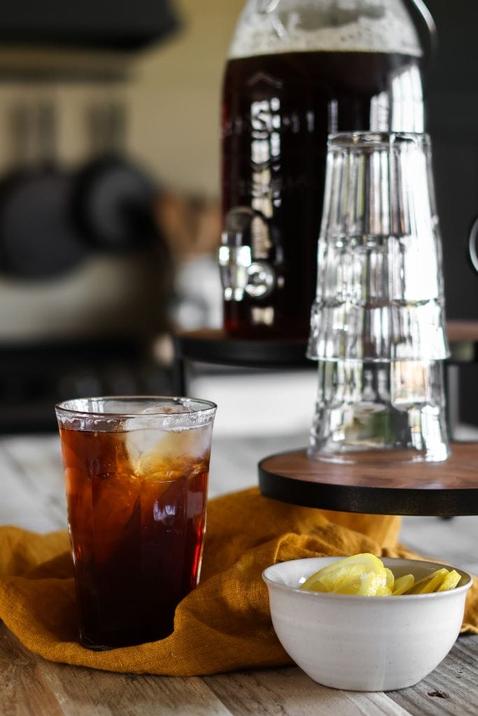 A tall glass pitcher filled with iced tea sits on a table with a bowl of lemon slices to place on the glass of ice tea.