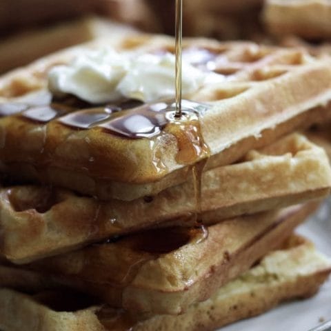 A close-up of a stack of three golden brown waffles, with a pat of melting butter on top and drizzled with syrup.