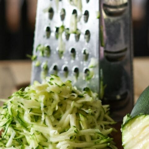 A box grater in the background with a pile of shredded zucchini laying on a cutting board