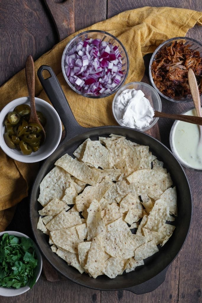 tortilla chips in a cast iron skillet with the toppings for bbq nachos around the skillet