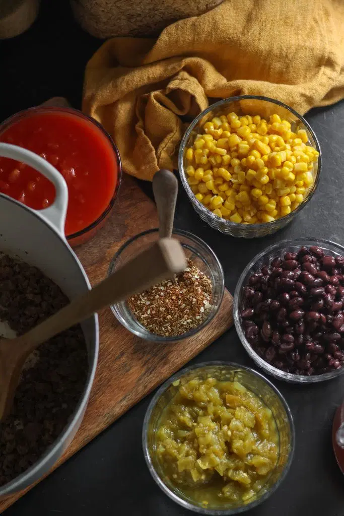 Ingredients for creamy taco soup including cans of diced tomatoes, diced green chilies, black beans, corn, cream cheese, and ground beef laid out on a kitchen counter.