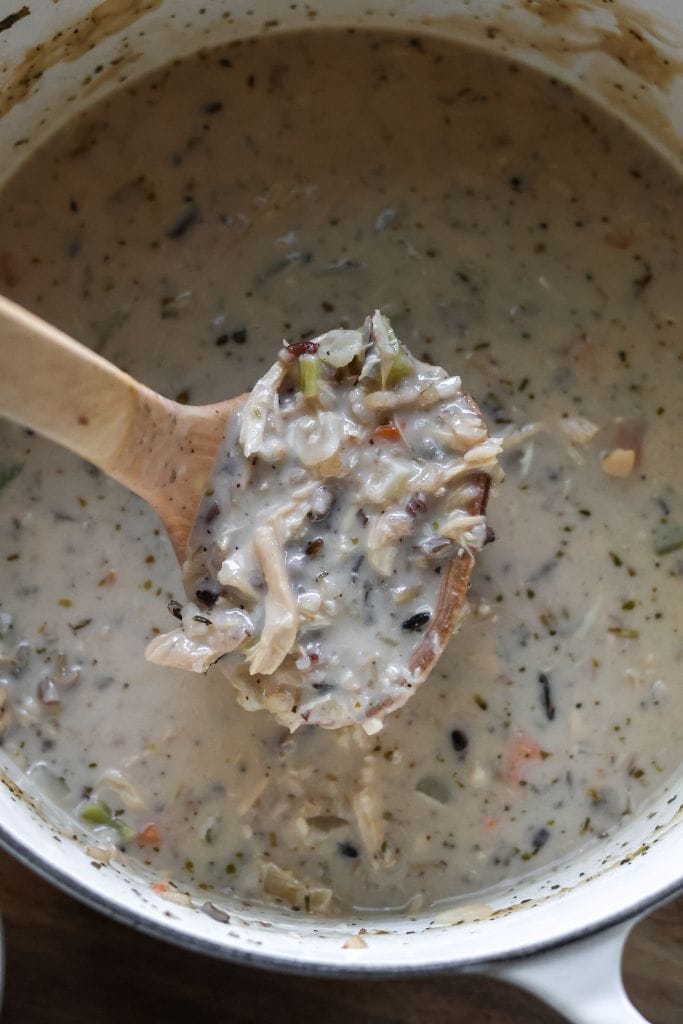 A ladle scooping creamy chicken and wild rice soup from a white Dutch oven, showcasing chunks of shredded chicken, vegetables, and wild rice in a rich broth.