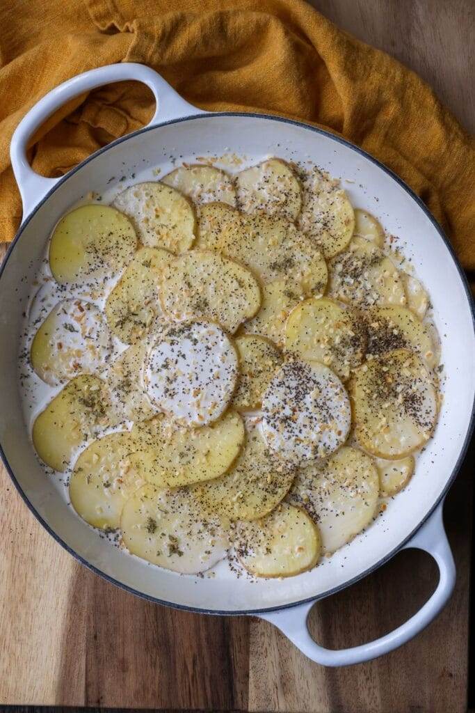 Layering thin slices of potatoes in a cast iron skillet, each layer seasoned with garlic, thyme, and cream, ready to be baked.