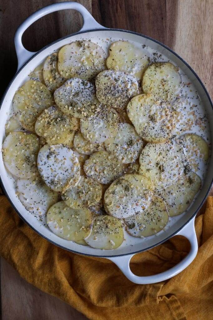 Close-up of thinly sliced potatoes being layered in a cast iron skillet, with herbs, garlic, and cream being poured over each layer.